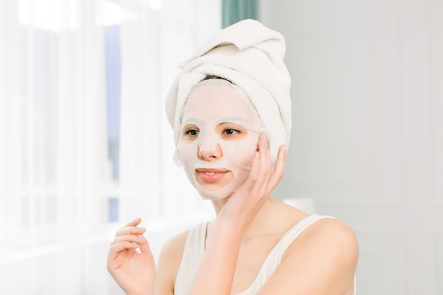 Closeup portrait de jeune jolie femme appliquant un masque de feuille de papier sur son visage. Procédure cosmétique. Spa de beauté et cosmétologie.