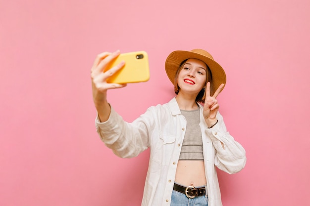 Closeup portrait de jolie fille en vêtements d'été et chapeau prend selfie sur fond rose pastel