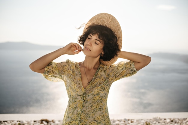 Closeup portrait of brunette curly lady sourit avec sensibilité les yeux fermés Femme aux cheveux courts en canotier et tenue florale pose sur fond de mer