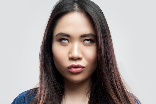 Closeup portrait of funny crazy belle brune asiatique jeune femme en veste en jean bleu décontracté avec maquillage debout avec les yeux croisés. tourné en studio intérieur, isolé sur fond gris clair.