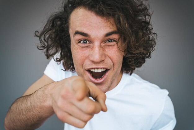 Closeup portrait en studio d'un homme de race blanche heureux et heureux porte des points de t-shirt blanc à vous choisit quelqu'un ou fait de la publicité pour un produit Bel homme souriant indique à la caméra isolée sur un mur gris