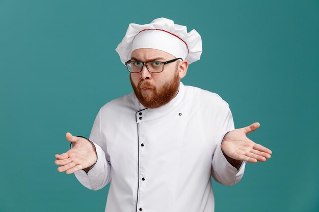 Clueless jeune chef masculin portant des lunettes uniformes et une casquette regardant la caméra montrant les mains vides isolées sur fond bleu