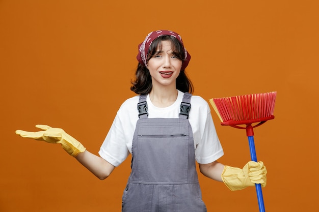 Clueless jeune femme nettoyeuse portant des gants en caoutchouc uniformes et un bandana tenant une vadrouille à raclette regardant la caméra montrant une main vide isolée sur fond orange