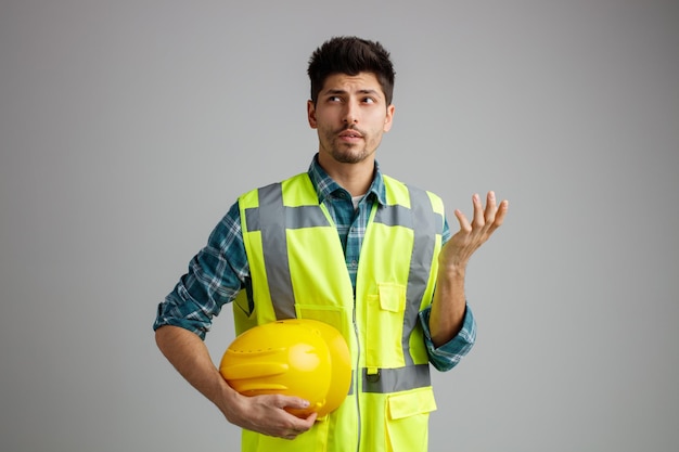 Clueless jeune ingénieur masculin portant l'uniforme tenant un casque de sécurité regardant le côté montrant la main vide isolé sur fond blanc