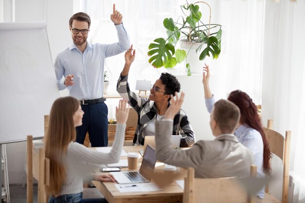Un coach d'affaires ou un mentor masculin du millénaire donne une formation ou une présentation de tableau à feuilles pour divers travailleurs des employés multiethniques se lèvent les mains engagées dans une activité de consolidation d'équipe remue-méninges au bureau