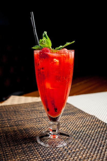 Cocktail alcoolisé à la fraise rouge à la menthe et à la paille. Cocktail alcoolisé ou non alcoolisé dans un verre clair long, haut et étroit avec un liquide rouge foncé et du sirop et de la menthe sur fond sombre