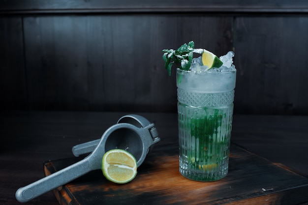 Cocktail alcoolisé "Mojito" avec de la glace et des feuilles de menthe fraîche avec des tranches de citron vert est sur la table à côté d'un outil professionnel pour obtenir du jus de citron ou de citron vert. Week-end au bar. Beau cocktail