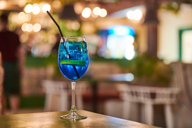 Cocktail Blue Lagoon en verre avec de la paille, espace de copie d'arrière-plan flou de lumière de café de nuit. Savoureux cocktail bleu sur la table du café. Liqueur bleue Curaçao avec vodka aromatisée et limonade