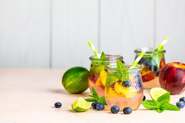 Cocktail d'eau infusé aux myrtilles et aux pêches, limonade ou thé en pot avec gouttes d'eau Boisson froide glacée d'été avec myrtille, citron vert, pêche et menthe