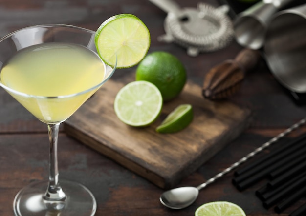 Cocktail Gimlet Kamikaze dans un verre à martini avec tranche de citron vert et glace sur fond de bois avec citrons verts frais et passoire avec shaker vue du dessus