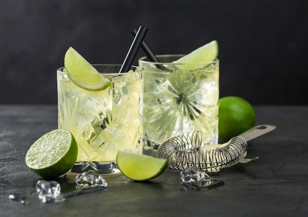 Cocktail Gimlet Kamikaze dans des verres en cristal avec une tranche de citron vert et de la glace sur une surface noire avec des limes fraîches et une passoire.