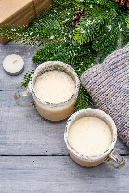 Cocktail de lait de poule dans une tasse à deux verres disposée avec décoration de Noël sur une table en bois clair. Branche de sapin à feuilles persistantes, cadeau, maillot confortable, neige artificielle