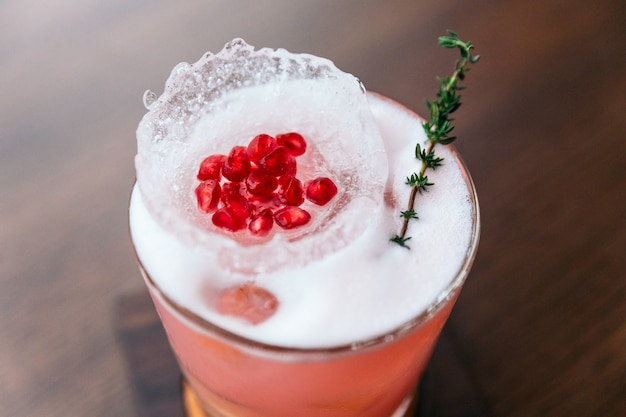 Cocktail rose grenade servi avec des graines dans un bol en forme de glace et du romarin.