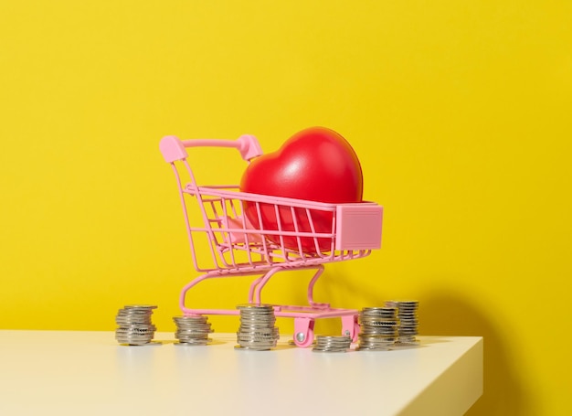 Coeur rouge dans un chariot en métal miniature du magasin et une pile de pièces sur fond jaune