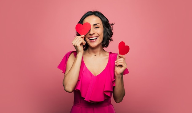 Coeur-yeux. Photo en gros plan d'une femme ravie vêtue d'une robe violette, qui tient deux cartes de la Saint-Valentin dans ses mains, se couvre les yeux d'une et rit.