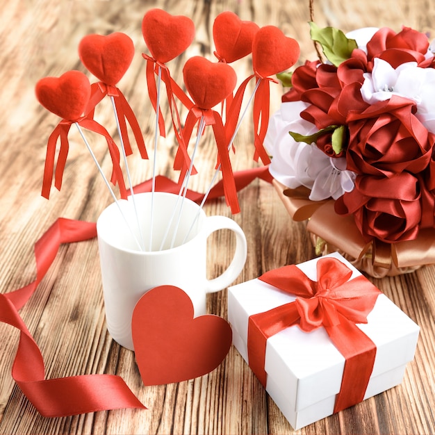 Coffret blanc avec noeud riibon rouge, tasse blanche avec coeurs rouges sur bâton, carte coeur et fleurs roses de ruban de satin sur fond en bois.