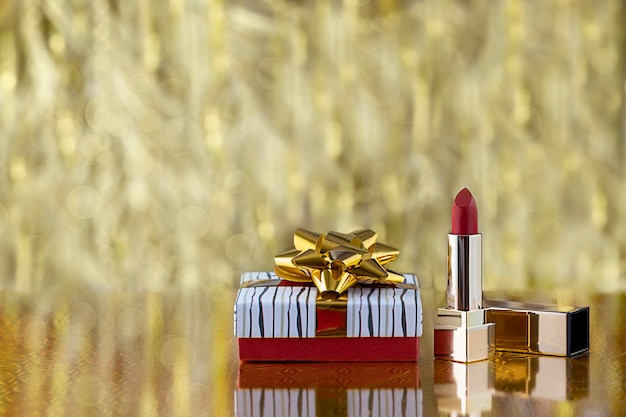 Photo coffret cadeau avec des arcs d'or et du rouge à lèvres rouge sur une pièce floue d'or