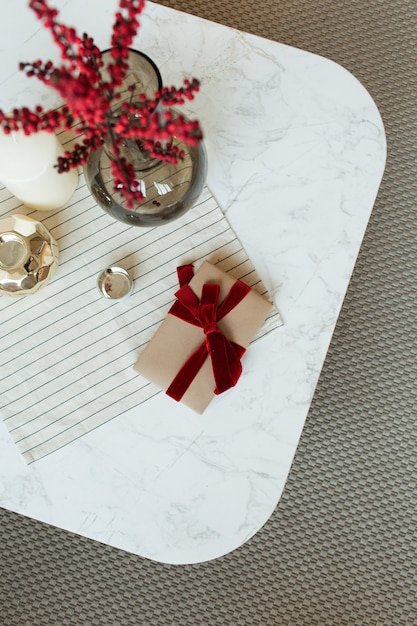 Coffret cadeau en papier fait main avec noeud papillon ruban rouge, baies rouges dans un vase en verre et décorations sur table en marbre