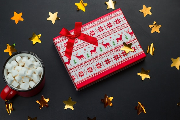 Coffret cadeau et tasse de guimauves sur fond noir avec des étoiles de confettis holographiques dorés