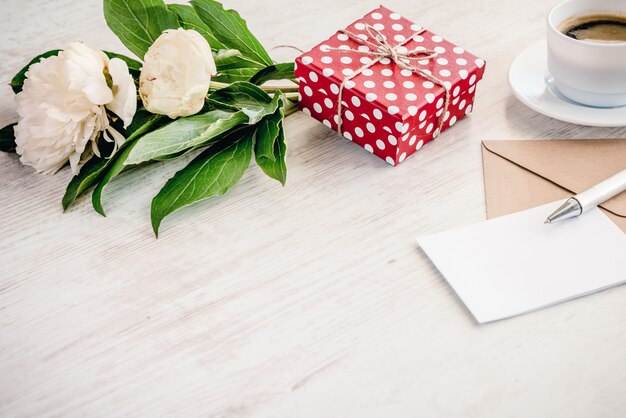 Coffret rouge en pointillé, enveloppe kraft et carte de voeux vide, bouquet de fleurs de pivoines et de tasses à café sur bois blanc, fond.