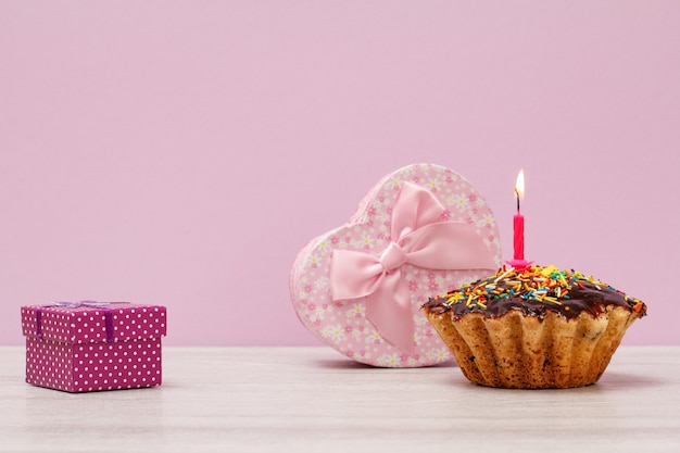 Coffrets cadeaux et muffin d'anniversaire savoureux avec glaçage au chocolat et caramel, décorés d'une bougie festive brûlante sur fond lilas. Concept minimal de joyeux anniversaire.
