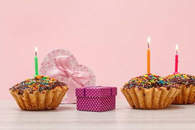 Coffrets cadeaux et muffins d'anniversaire savoureux avec glaçage au chocolat et caramel, décorés de bougies festives allumées sur fond bois et rose. Notion de joyeux anniversaire.