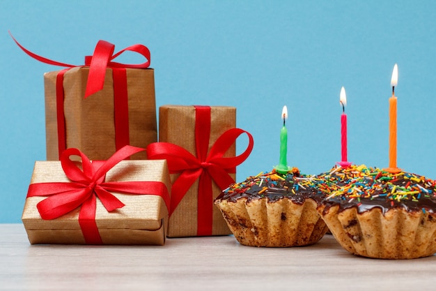 Coffrets cadeaux et trois délicieux cupcakes d'anniversaire avec glaçage au chocolat et caramel, décorés de bougies festives allumées sur fond bleu. Concept minimal de joyeux anniversaire.