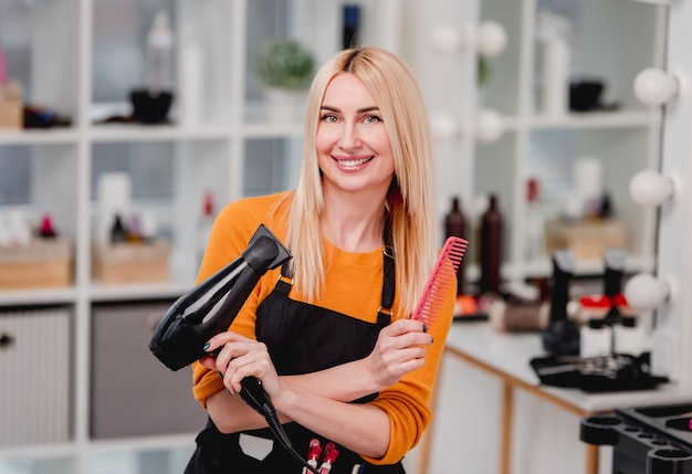 Coiffeur de belle fille blonde regardant la caméra, souriant et tenant un sèche-cheveux au salon de beauté