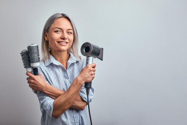 Coiffeur blonde jolie femme avec un outil professionnel posant sur fond de caméra, gris.