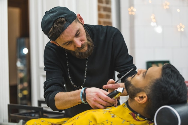 Coiffeur coupant la barbe d'un client à l'aide d'une tondeuse portative dans un salon de coiffure moderne