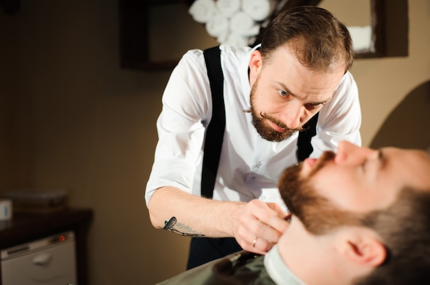 Le coiffeur coupe les cheveux et la barbe de l'homme dans le salon de coiffure