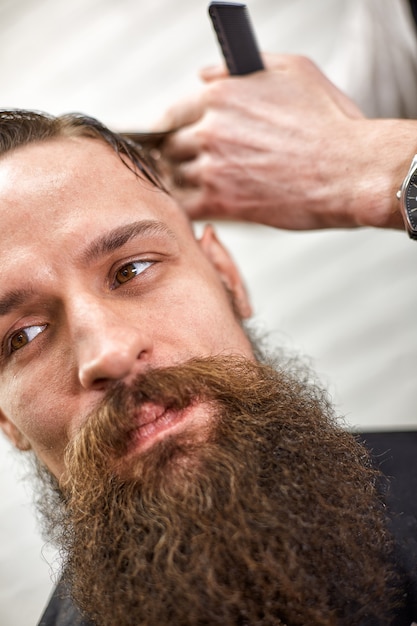 Photo un coiffeur élégant coupe un homme brutal avec une barbe épaisse. salon de coiffure léger. les hommes font une coupe de cheveux à la mode.