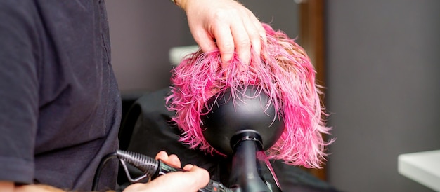 Coiffeur faisant une coiffure à l'aide d'un sèche-cheveux soufflant sur des cheveux roses personnalisés dans un salon de beauté.