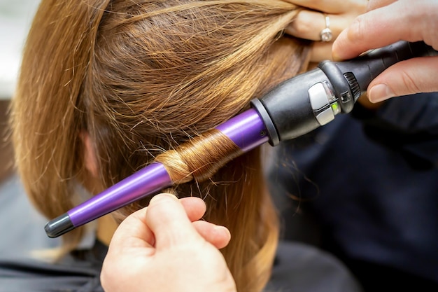 Le coiffeur fait des boucles de coiffure de longs cheveux bruns avec le fer à friser dans le salon de coiffure en gros plan