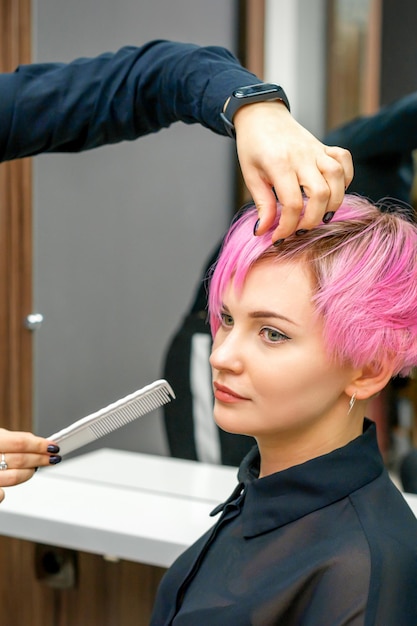 Coiffeur féminin coiffant les cheveux roses courts de la jeune femme blanche avec les mains et le peigne dans un salon de coiffure.