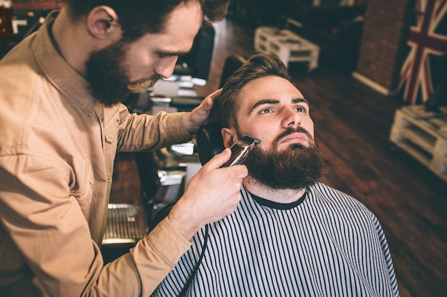 Photo un coiffeur gentil et bon coupe une partie de la barbe du client avec un rasoir électrique. il le fait très précisément.