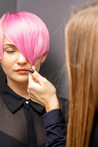 Le coiffeur avec les mains vérifie et fixe la courte coiffure rose de la jeune femme blanche dans un salon de coiffure