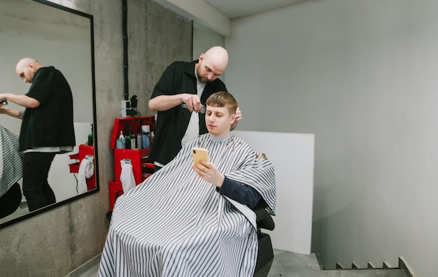 Coiffeur masculin coupe un jeune homme à l'aide de son smartphone dans une chaise de coiffeur
