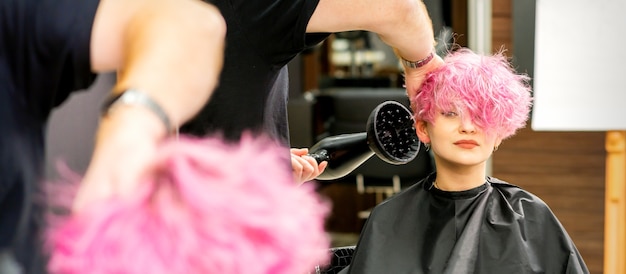 Un coiffeur masculin séchant les cheveux roses élégants de la cliente avec un sèche-cheveux dans un salon de beauté