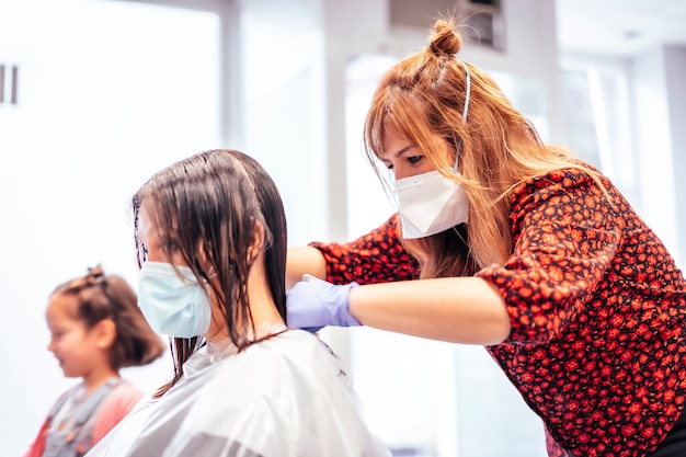 Coiffeur avec masque et gants coupant un client et la fille du client jouant ailée. Réouverture avec des mesures de sécurité pour les coiffeurs dans la pandémie de Covid-19