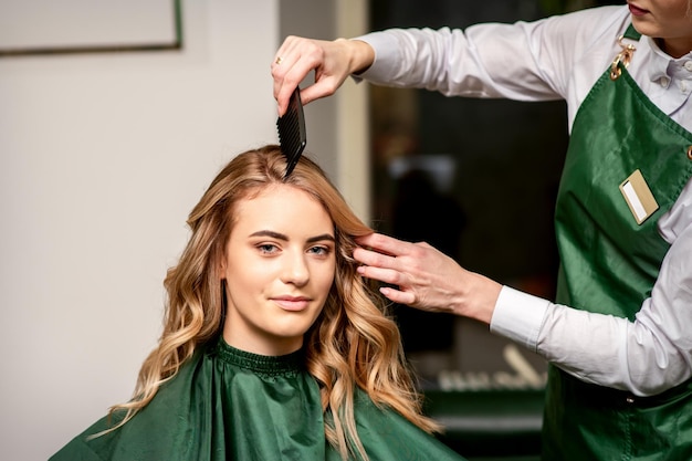 Coiffeur peignant les cheveux longs d'une jeune femme caucasienne regardant la caméra et souriant dans un salon de beauté