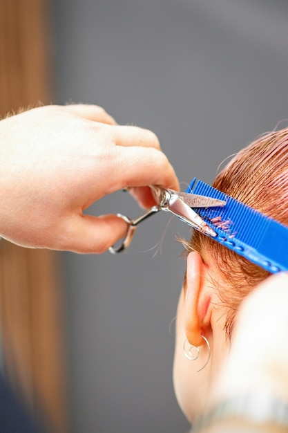 Un coiffeur professionnel coupe des cheveux roses courts avec des ciseaux dans un salon de coiffure en gros plan.
