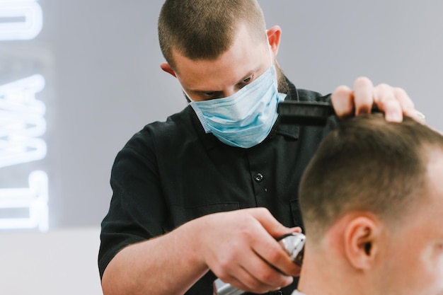 Coiffeur Professionnel Dans Un Masque De Gaze Médicale Sur Le Visage Coupe Les Cheveux Du Client Avec Une Tondeuse Avec Un Regard Concentré Le Coiffeur Travaille En Quarantaine Gros Plan