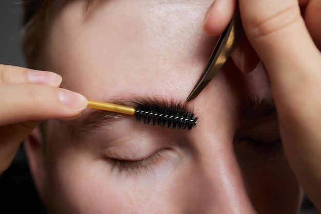 Coiffeur professionnel faisant la procédure de filetage et corrigeant la forme des sourcils au jeune client masculin avec une pince à épiler dans le salon de coiffure Coiffeur au travail Concept de soins des sourcils Plumer les sourcils