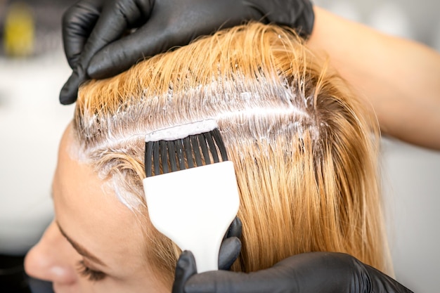 Le coiffeur teint les racines des cheveux blonds avec une brosse pour une jeune femme dans un salon de coiffure