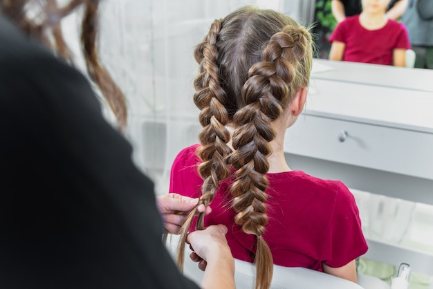 Coiffeur tisse une tresse à une fille blonde préadolescente dans un salon de beauté et de coiffure