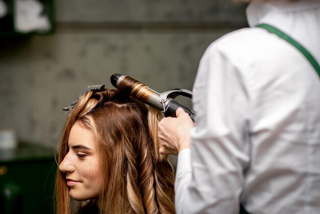 La coiffeuse frise les cheveux d'une jeune femme caucasienne aux cheveux bruns dans un salon de beauté