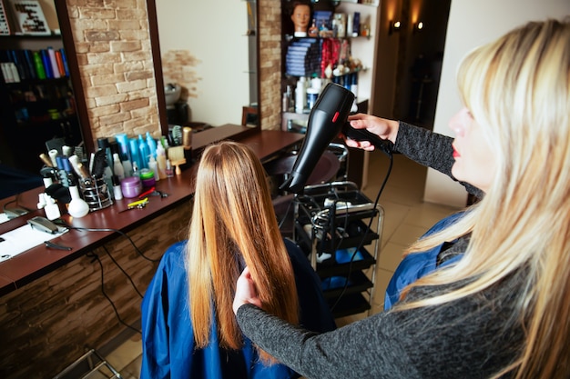 Coiffeuse professionnelle faisant la coiffure à l'aide d'un sèche-cheveux pour jeune femme dans un salon de beauté.