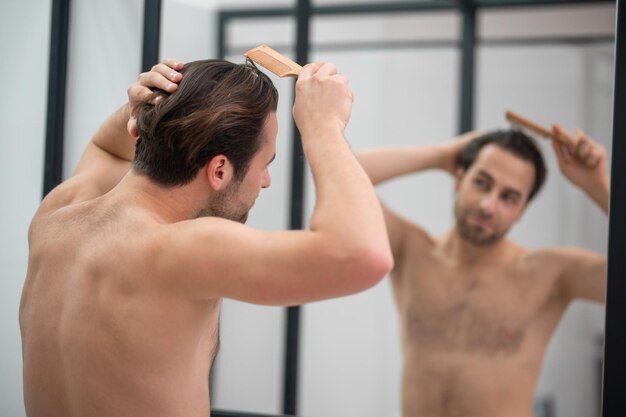 Coiffure. Jeune bel homme se brossant les cheveux et ayant l'air satisfait