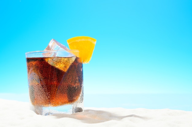 Cola rafraîchissant avec de la glace et une tranche d'orange dans un verre sur la côte de sable